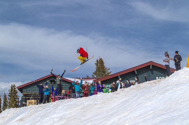 A skiier in a bright red jacket mid-jump, with a cheering crowd and lodgein the background
