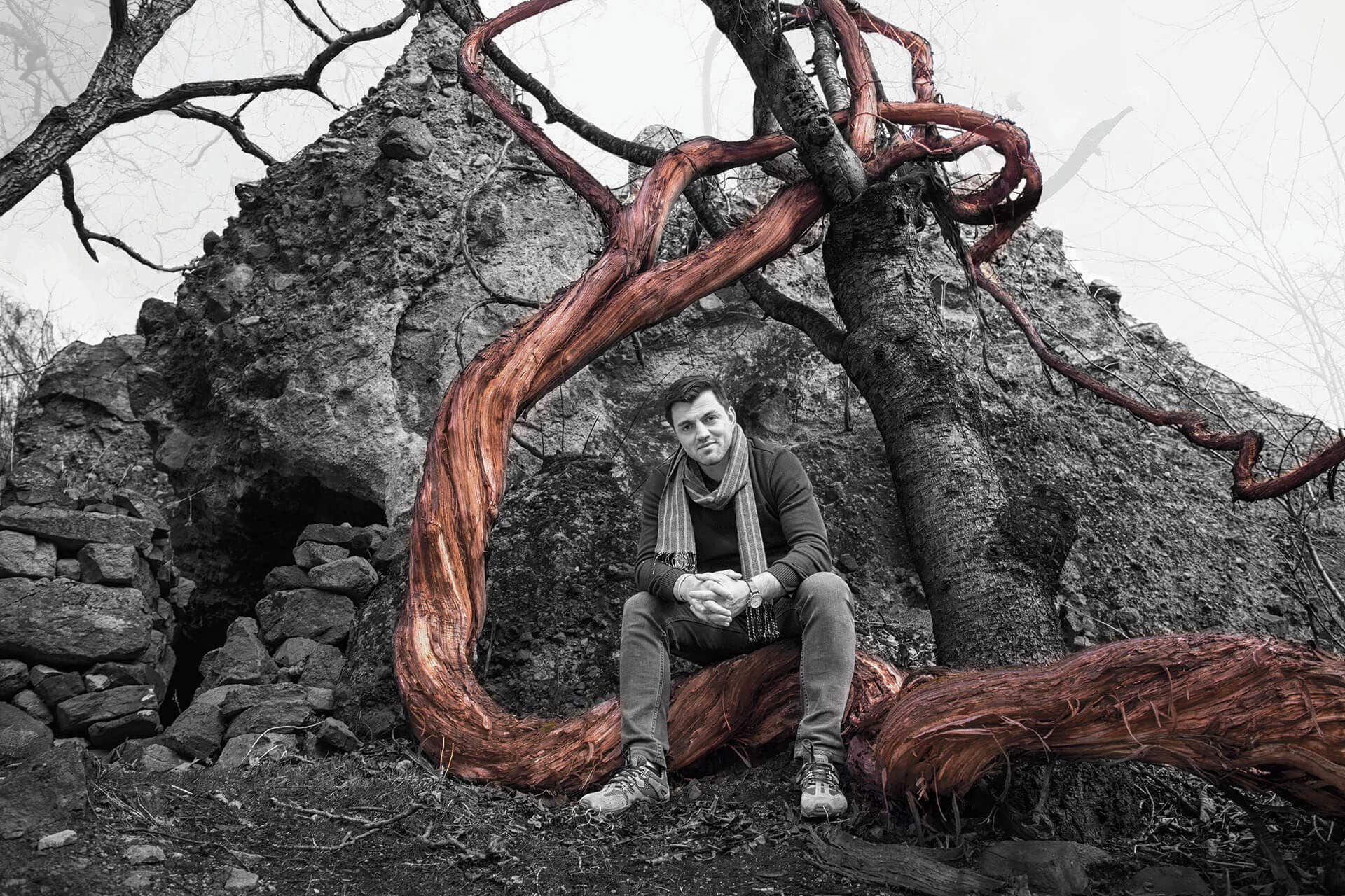 Black and white photo of man sitting on a huge vine in front of a rocky cave-like hill