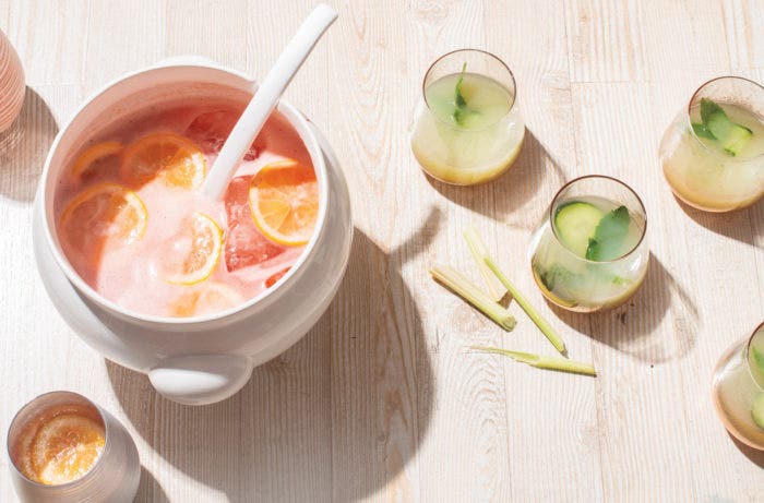 Large white punchbowl with pink drink and lemon slices, a cup with that drink and a lemon slice, three glasses of yellow-green drink with cucumber slices and sage leaves in it