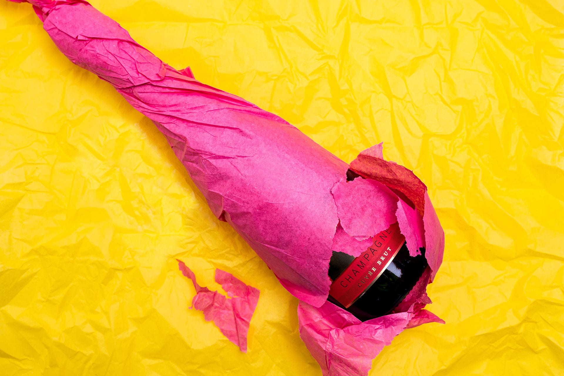 Champagne bottle wrapped in hot pink tissue paper against a bright yellow background